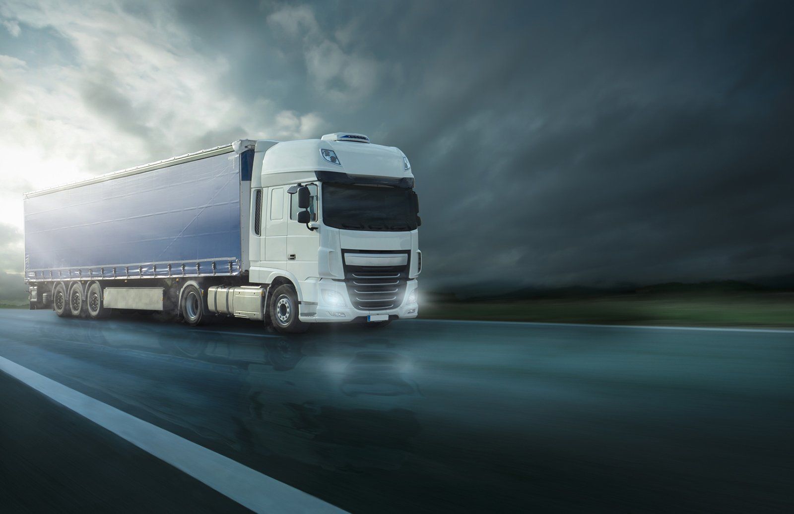 A white semi-truck with a blue trailer drives on a wet road under a dark, stormy sky.