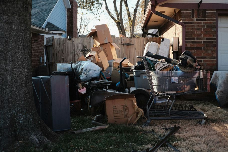 Piled-up cardboard, bags, and discarded furniture clutter a narrow yard beside a brick house.