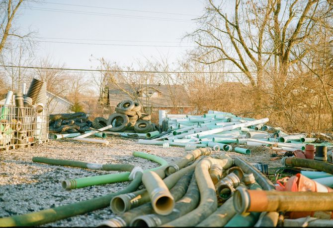 Pile of discarded pipes and scrap metal in an overgrown yard with trees and a small building in the background