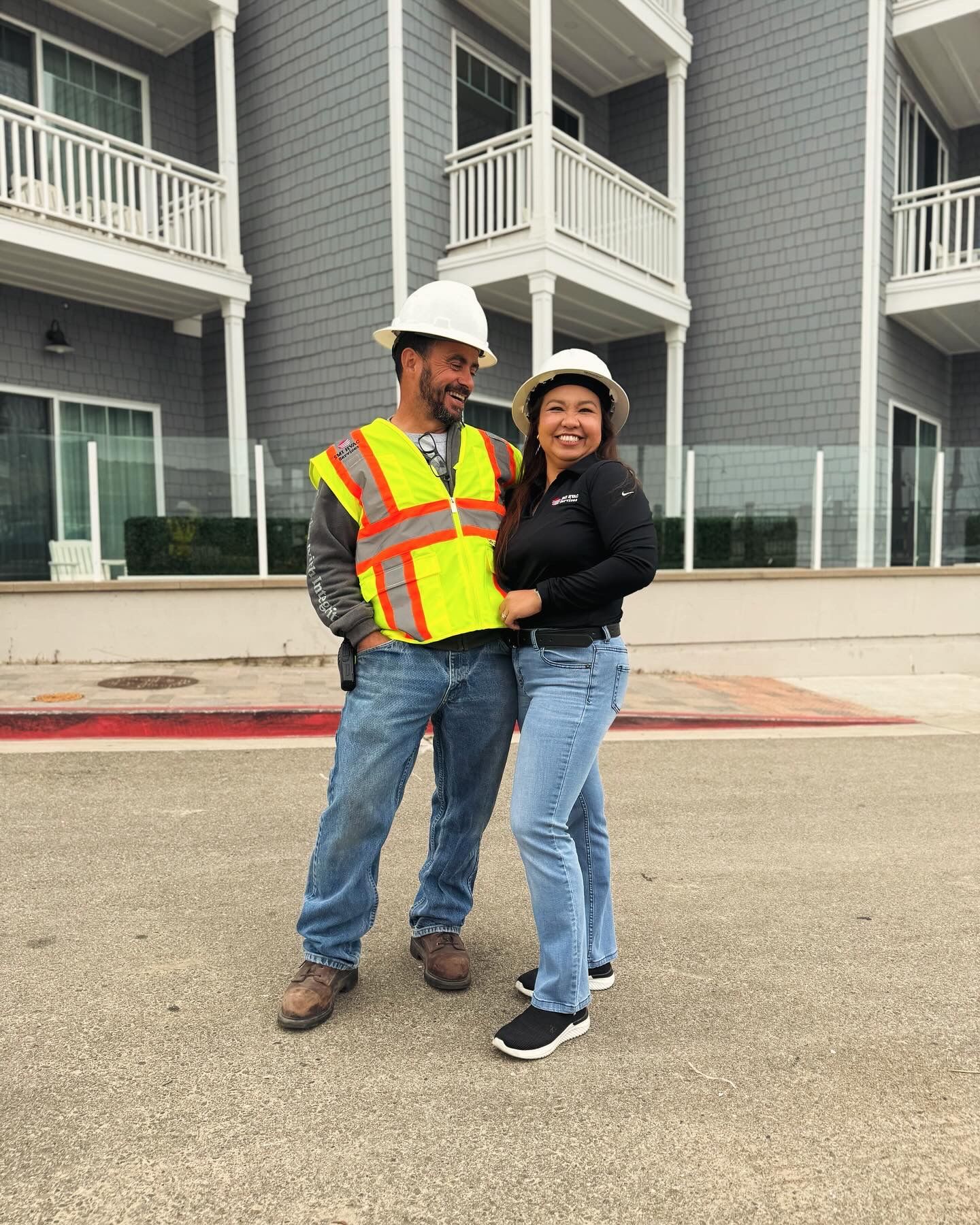 Two people in construction gear smile in front of a gray building.