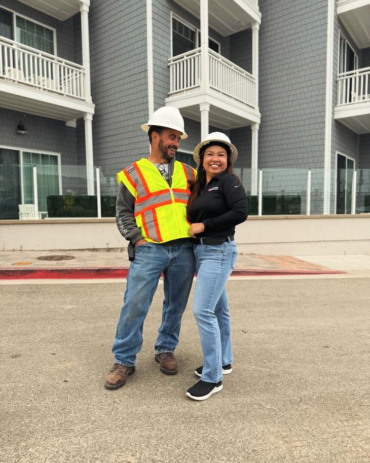 Two people in construction gear smile in front of a gray building.