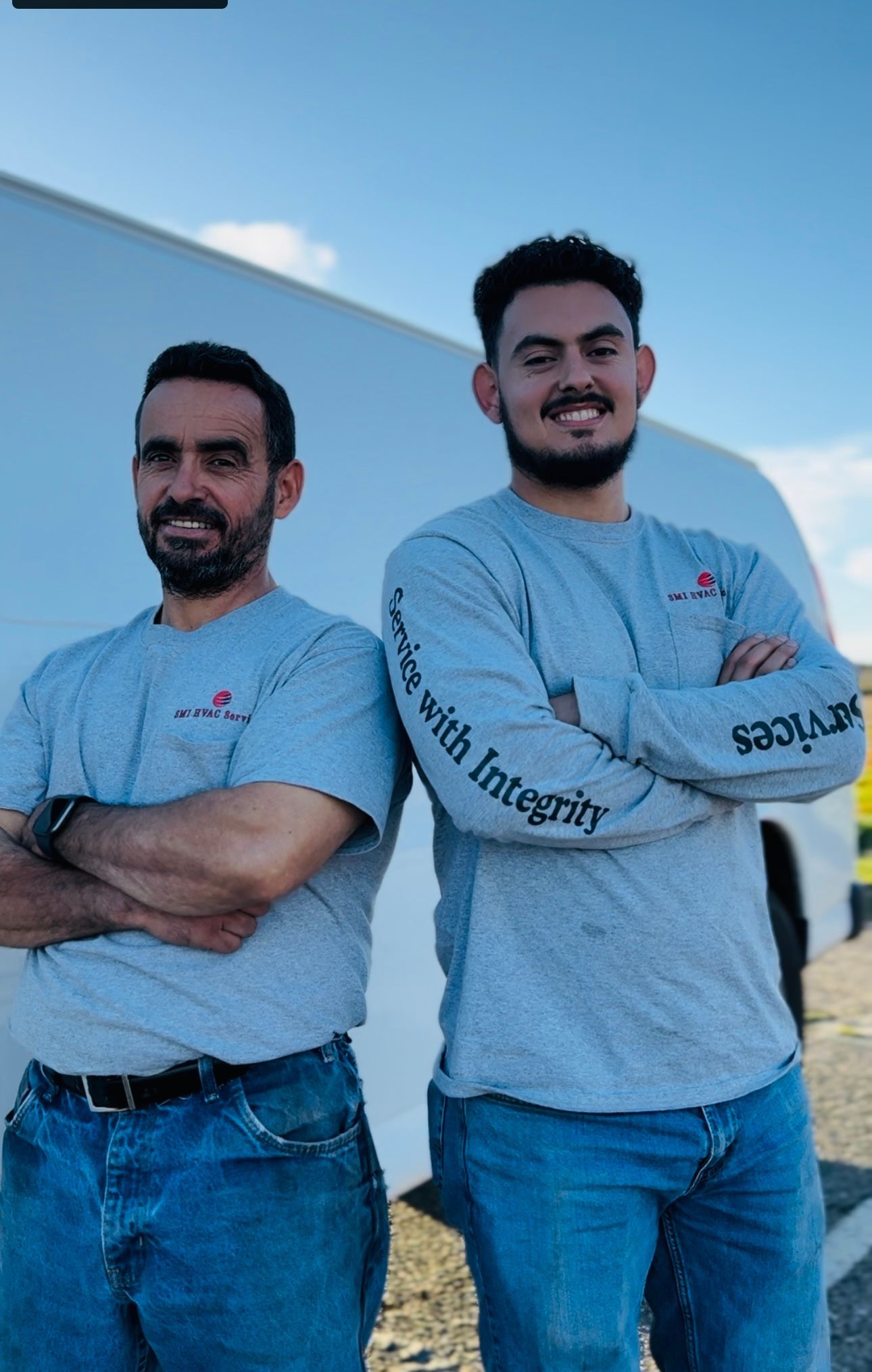 Two men in gray shirts and jeans, arms crossed, standing in front of a trailer.