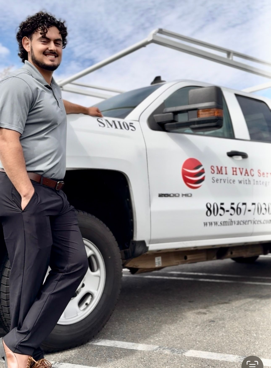 Man in gray shirt and black pants leans against a white HVAC service truck.