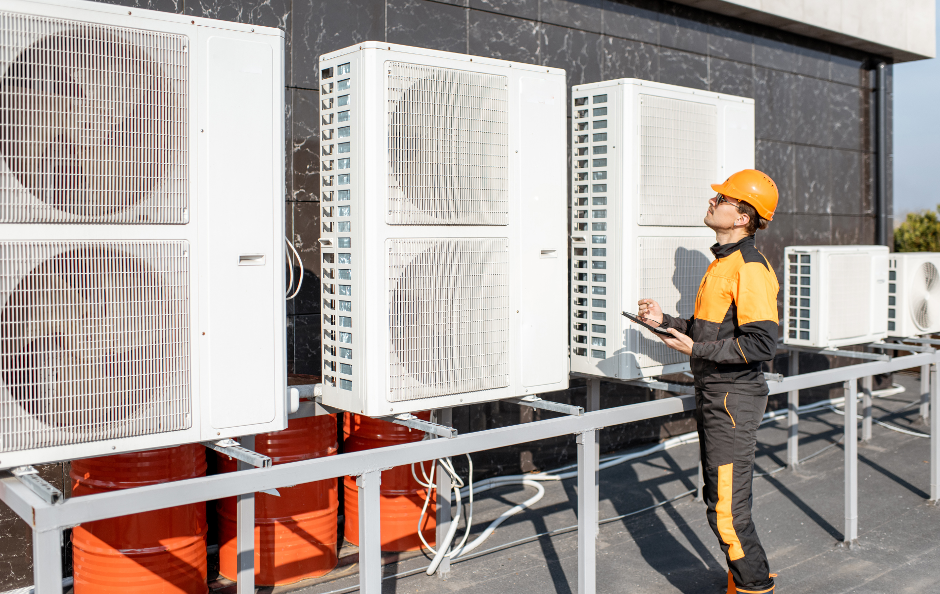 HVAC technician in orange hardhat inspects rooftop air conditioning units, holding a tablet.