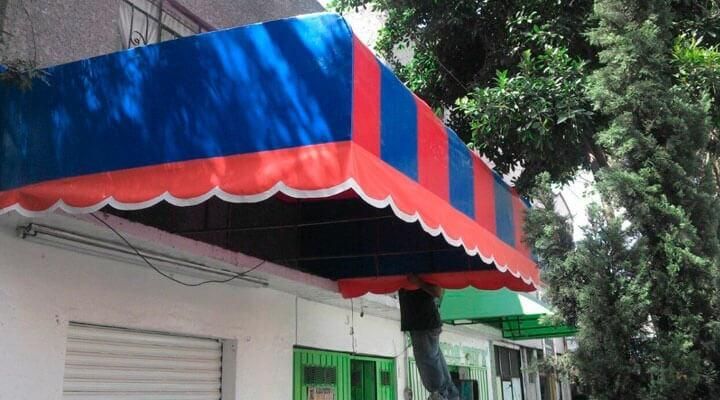 Un hombre está parado bajo un toldo azul y rojo al costado de un edificio.