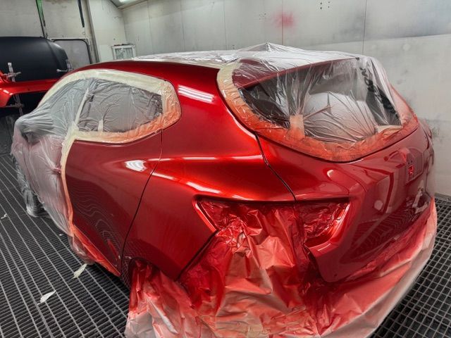 In uno stabilimento industriale, un braccio robotico montato su una struttura blu spruzza vernice rossa sul cofano scuro di un'auto.