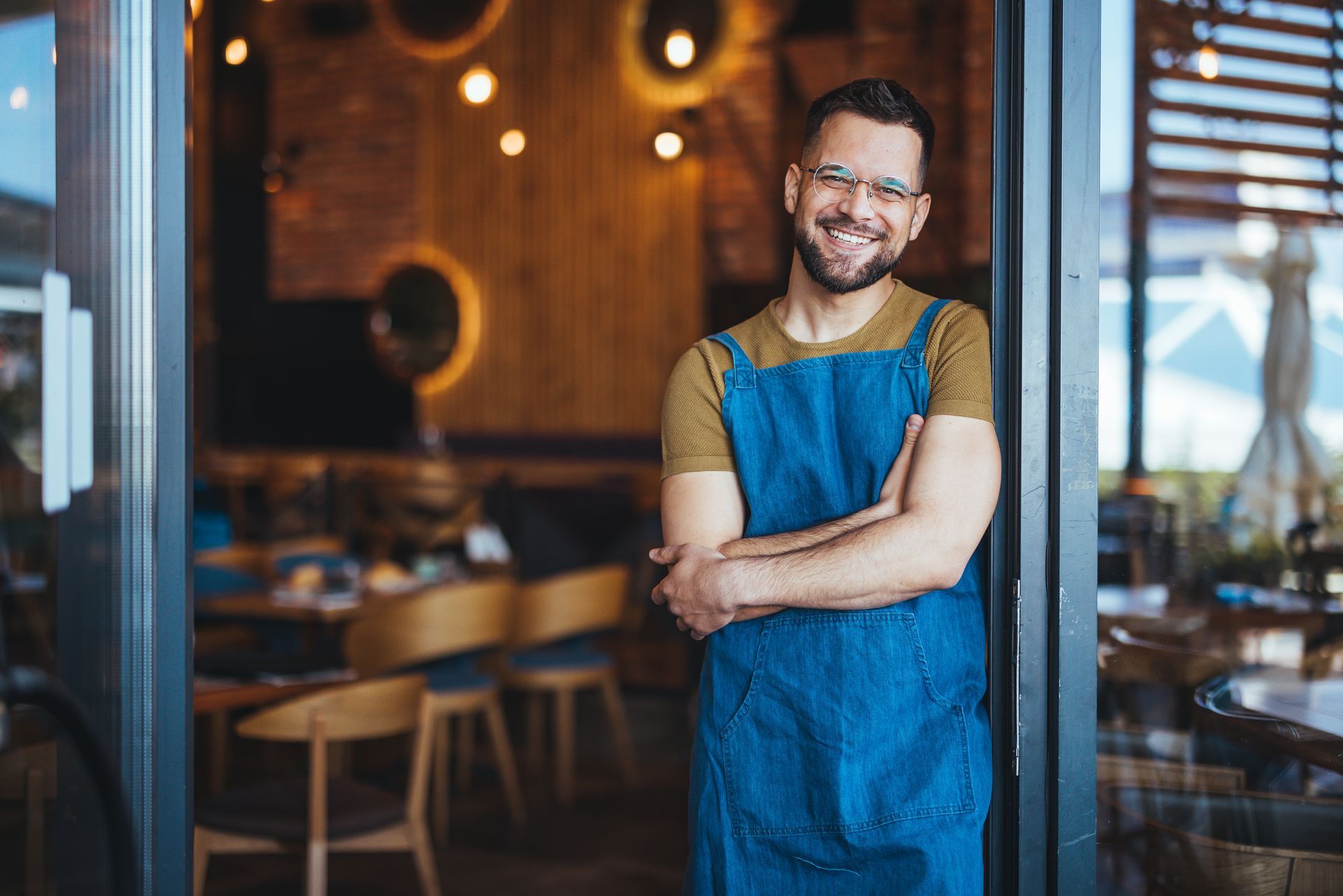 A smiling person wearing a tan t-shirt and blue apron stands with arms crossed in the doorway of a dimly lit restaurant.