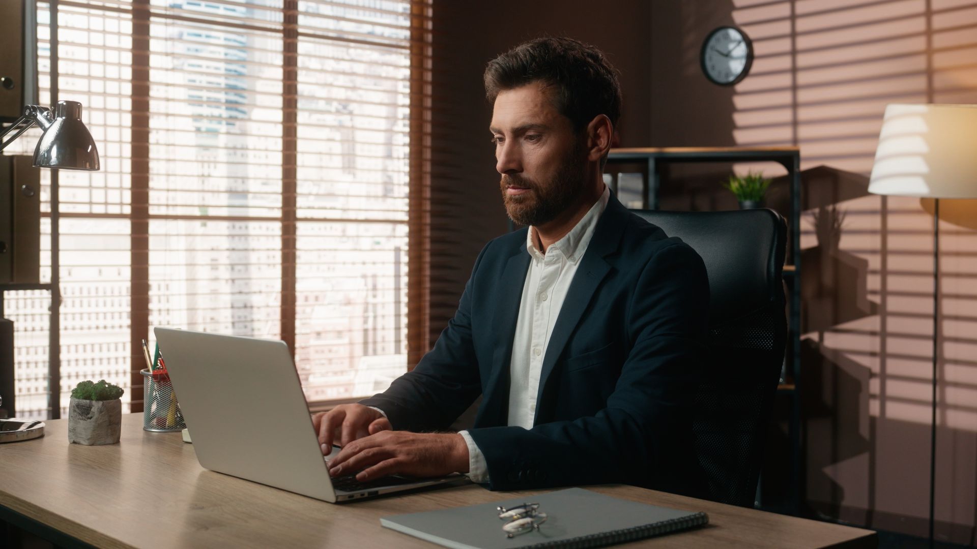 A focused person in a navy blazer types on a laptop at a desk in a sunlit home office with window blinds.