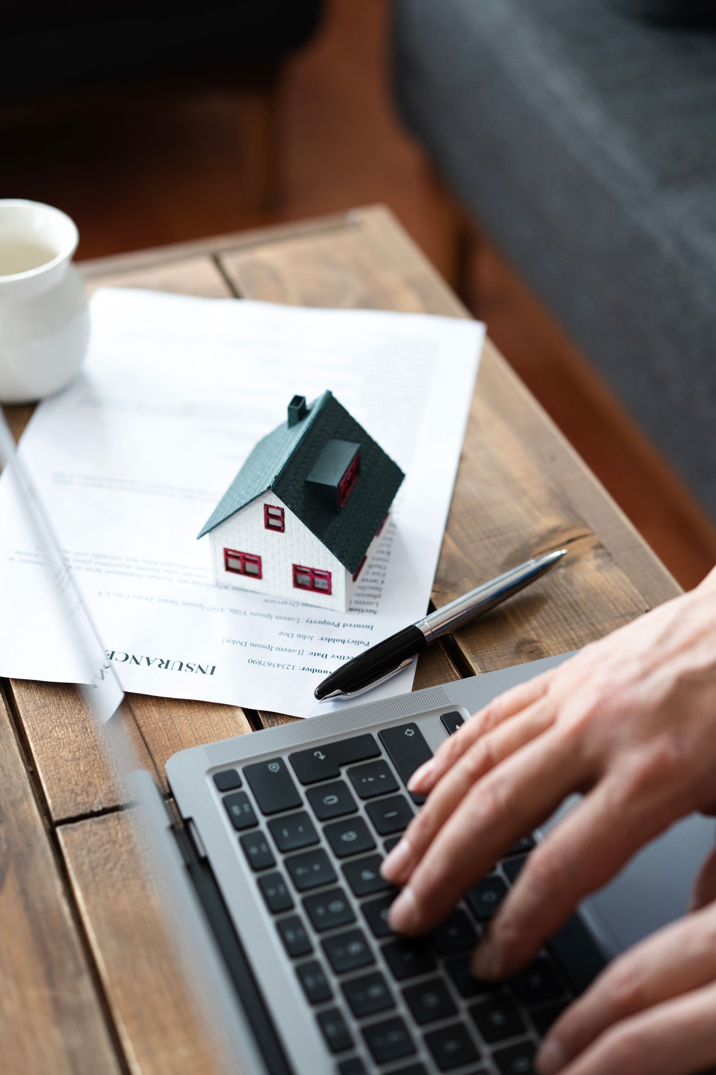 A home insurance agent is working on a laptop with a house model and an insurance policy on a desk.
