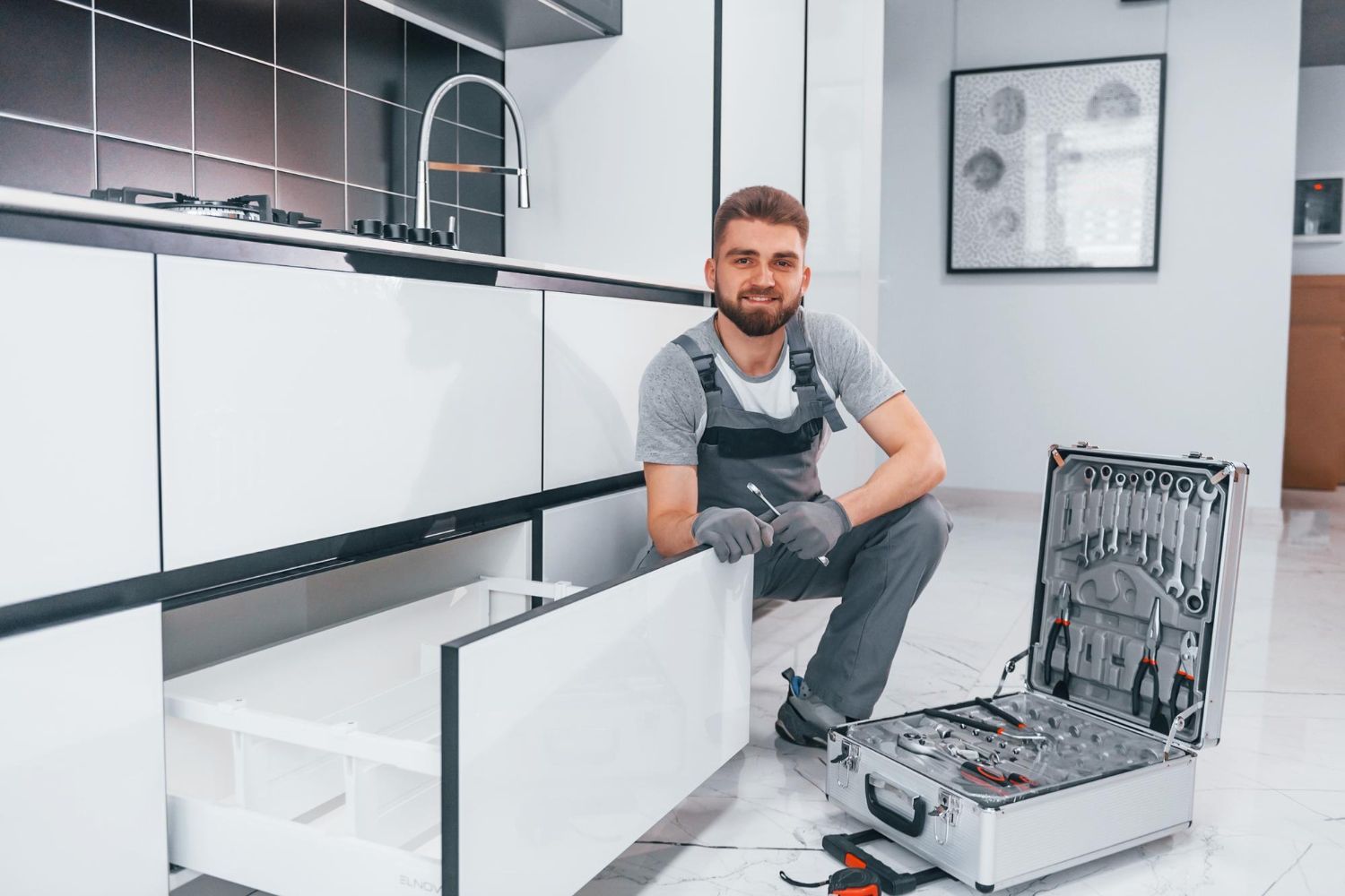 A man is kneeling on the floor in a kitchen next to a suitcase filled with tools.