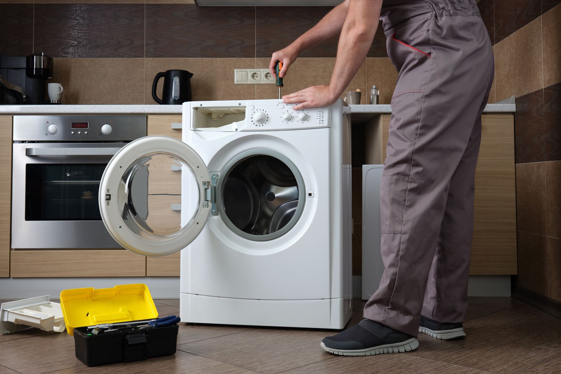A person repairs a washing machine in a kitchen, using a screwdriver. Tools sit on the floor.