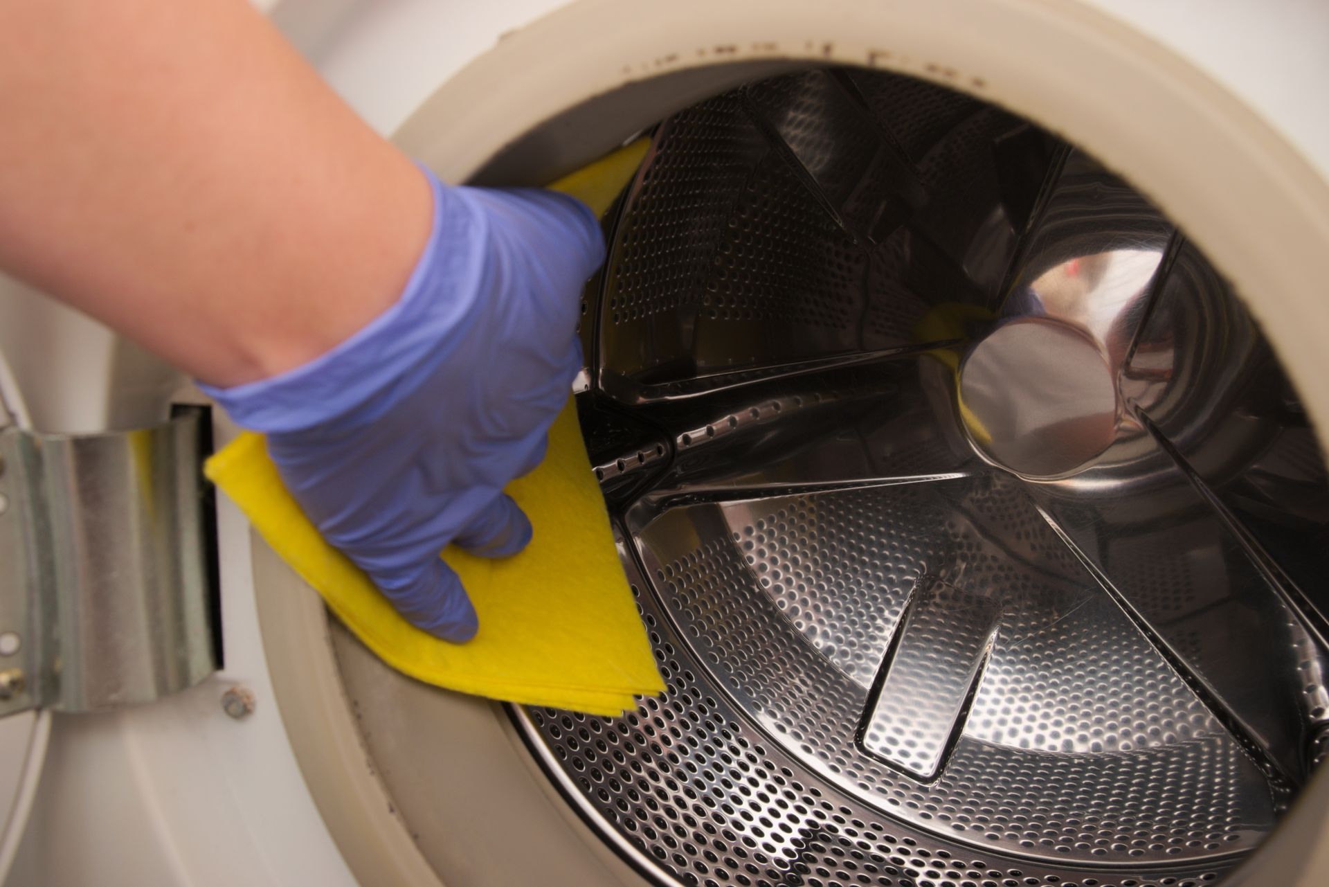 A gloved hand cleans the inside of a washing machine with a yellow sponge.