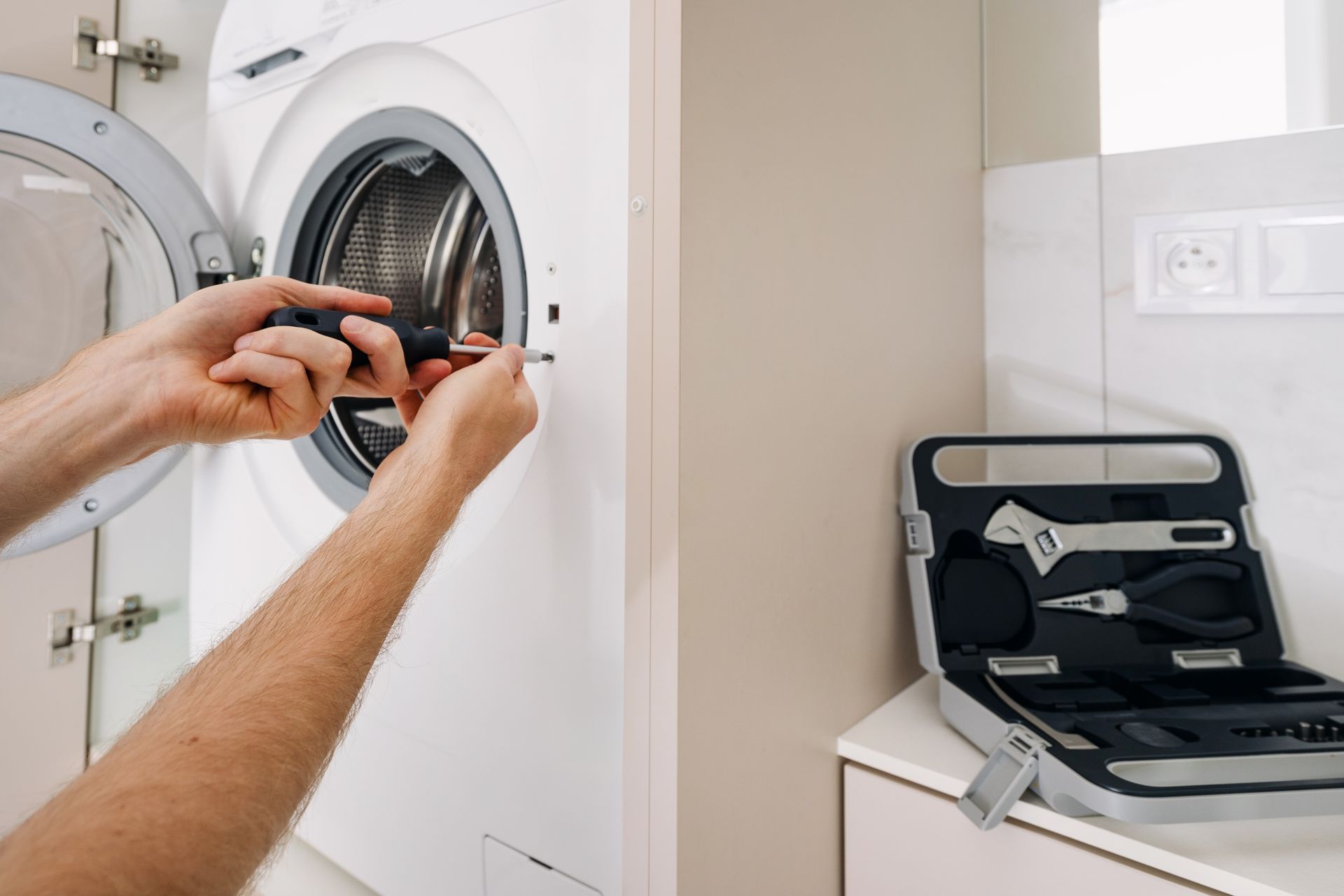 Person repairs a washing machine with a screwdriver; a tool kit sits nearby.
