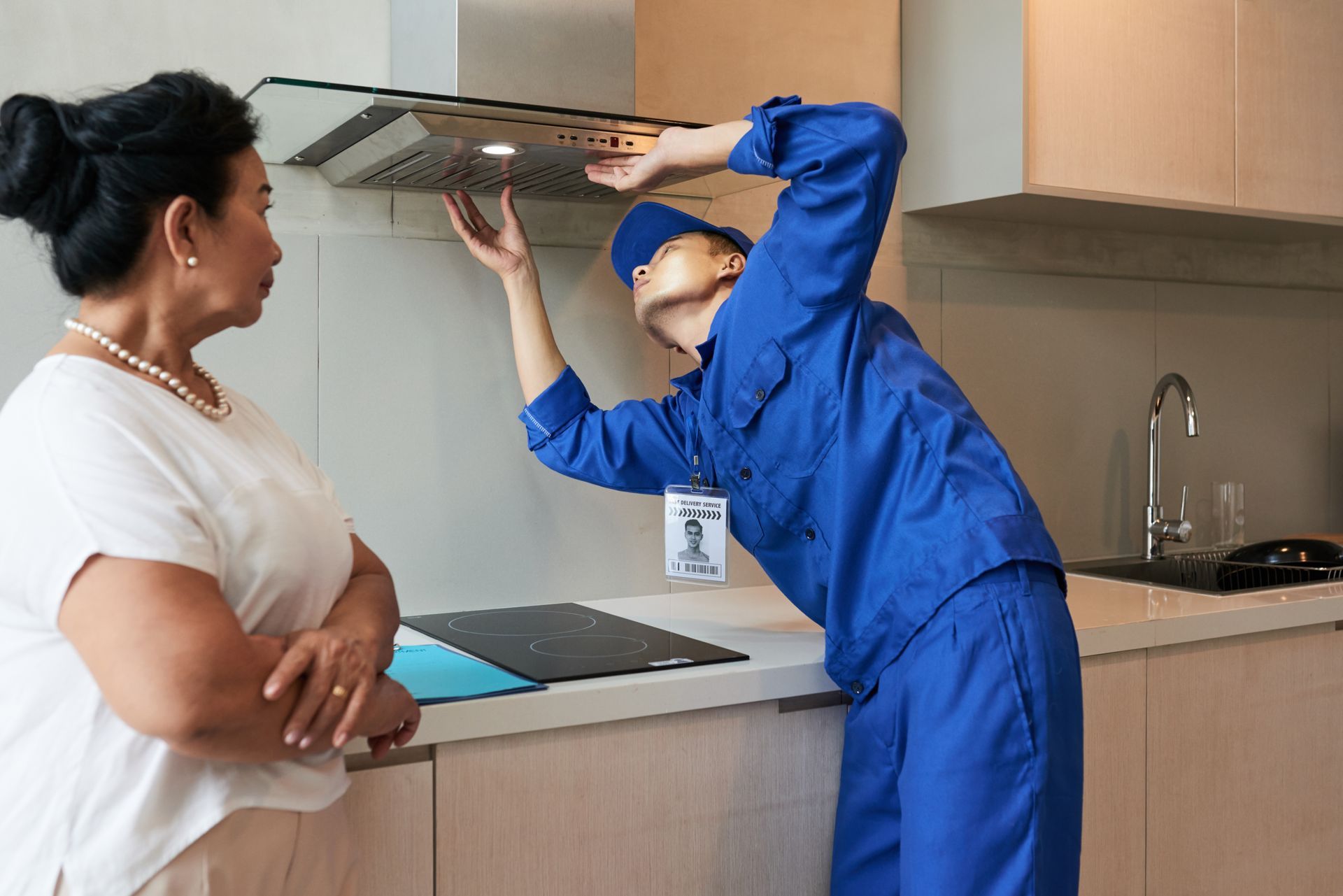 A repair person in blue uniform fixing a kitchen hood as a woman watches.