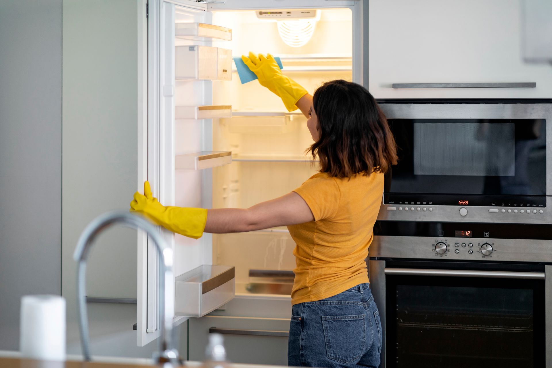 Woman wearing yellow gloves cleaning the inside of a refrigerator with a blue sponge in a kitchen.