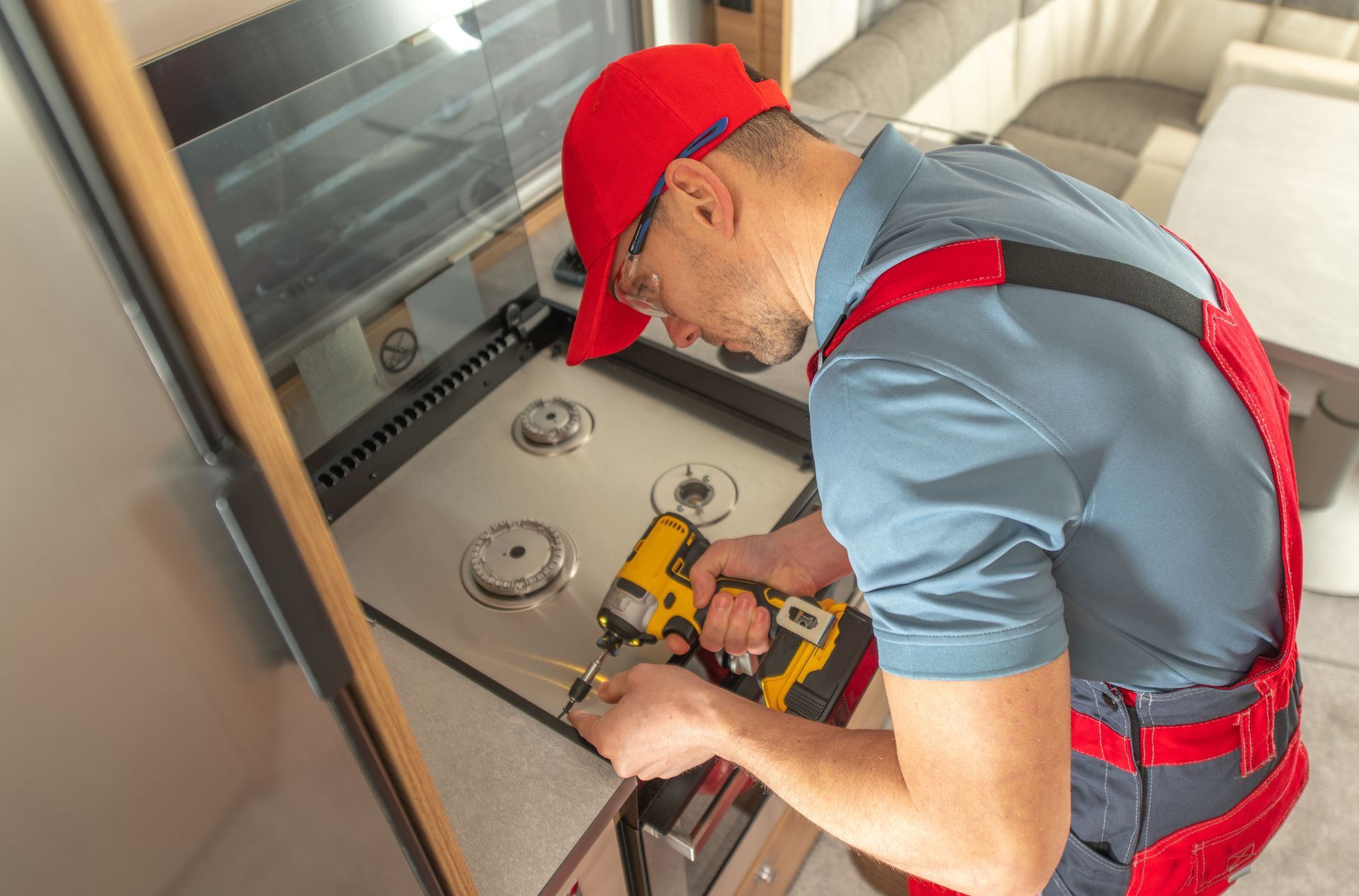 Person in red overalls and hat using a drill on a stove inside a recreational vehicle.