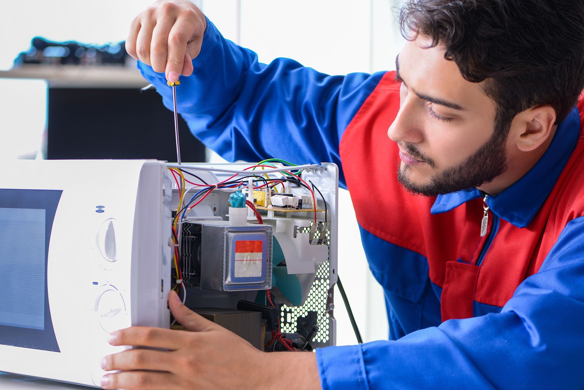 Man in blue and red uniform repairing a white microwave using a screwdriver. Indoors.