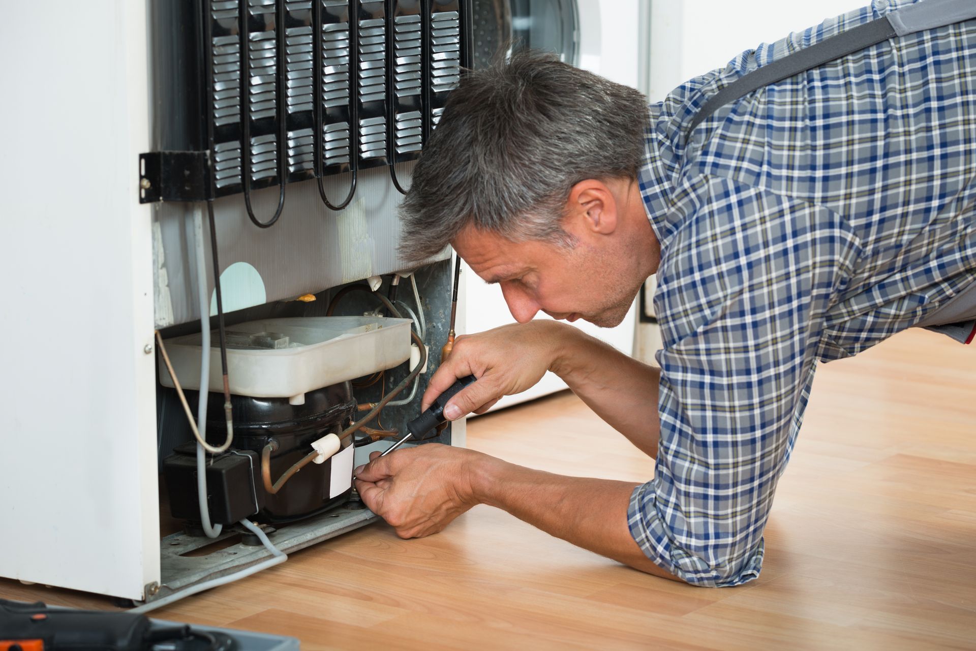 A man is kneeling on the floor fixing a refrigerator.