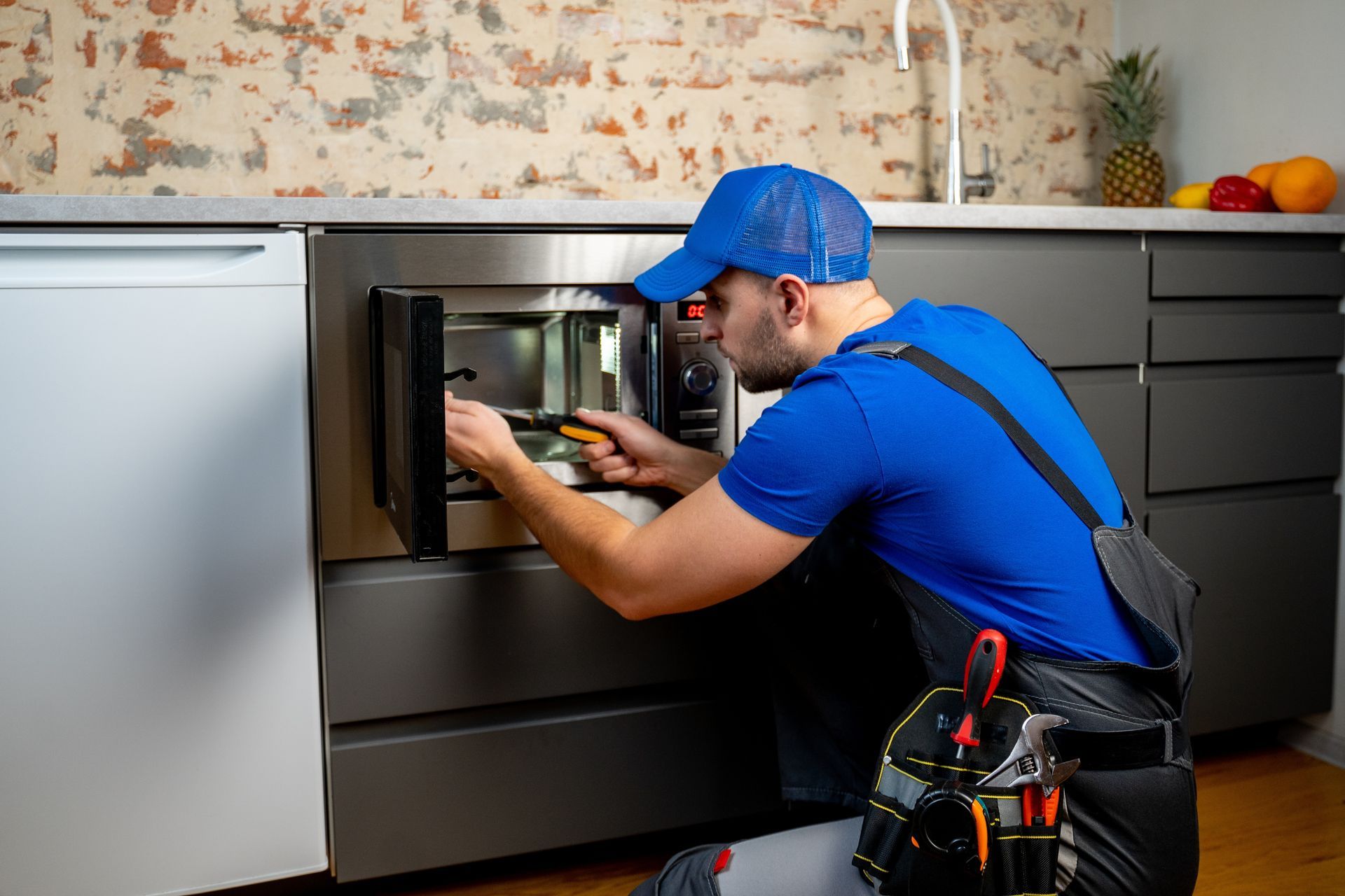 A person in blue uniform repairing a microwave in a kitchen.