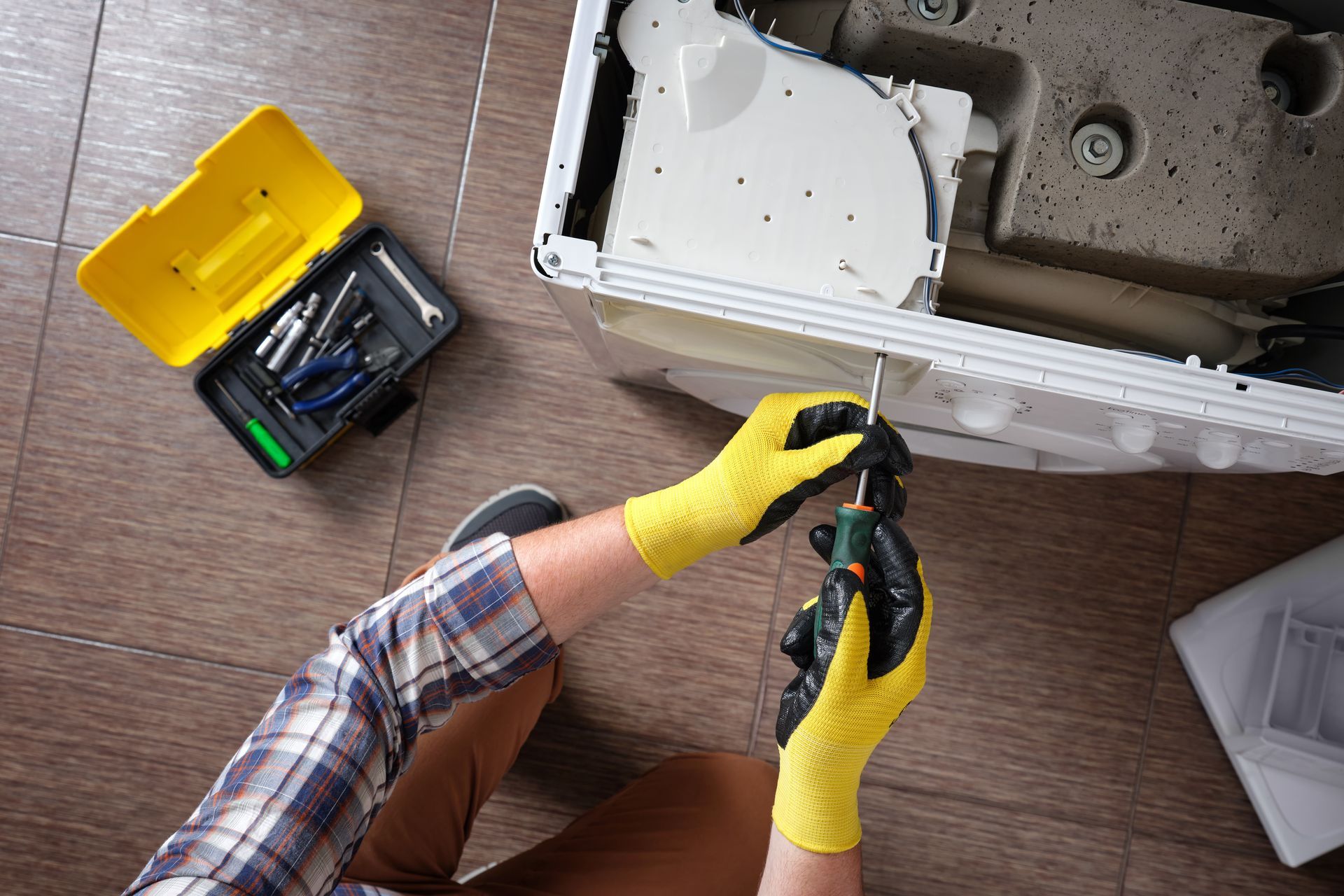Person in yellow gloves repairs appliance with a screwdriver, with a toolbox nearby.