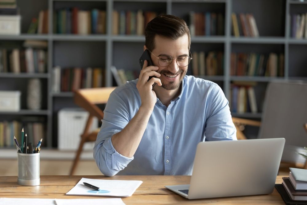 A Man Is Sitting At A Desk With A Laptop And Talking On A Cell Phone — Kaplan Engineering Pty Ltd In Bowen, QLD