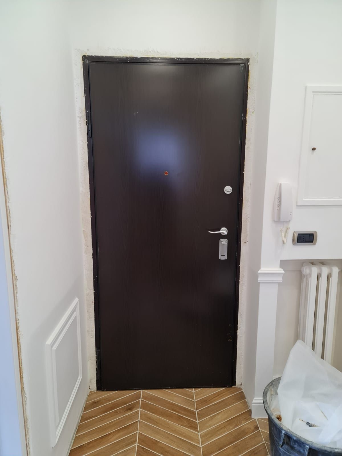 Porta marrone in un corridoio con pavimento in legno a spina di pesce e pareti bianche.