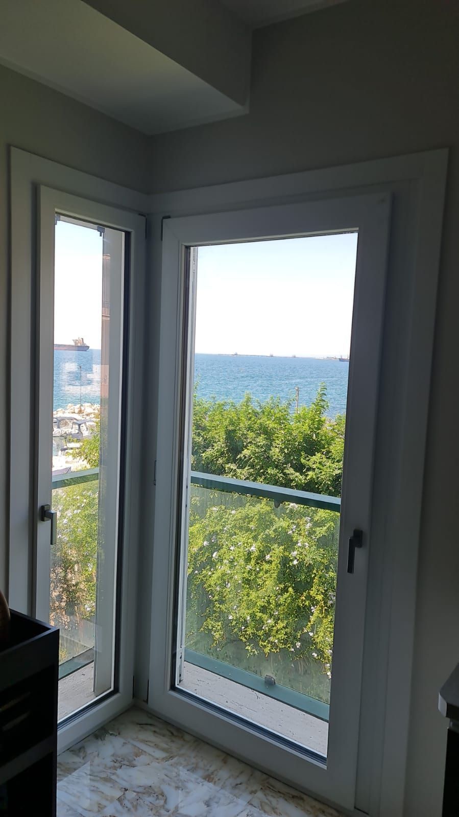 Finestra angolare con vista mare; sono visibili alberi e balcone.