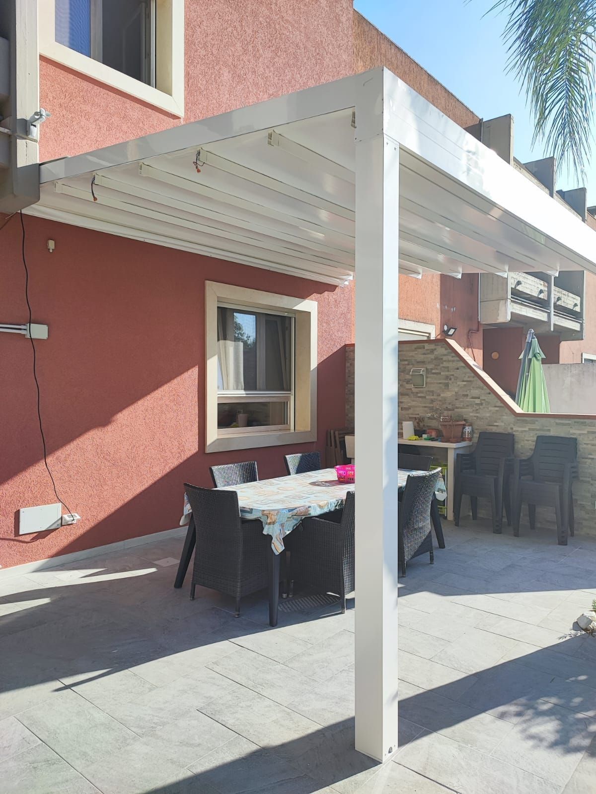 Pergola bianca sopra il tavolo da pranzo e le sedie all'aperto, contro una parete in stucco rosso.
