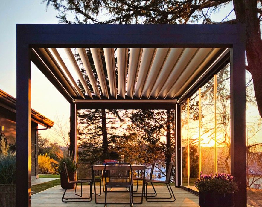 Pergola con tetto a doghe regolabili sopra il tavolo da pranzo all'aperto, vista del tramonto attraverso le finestre.