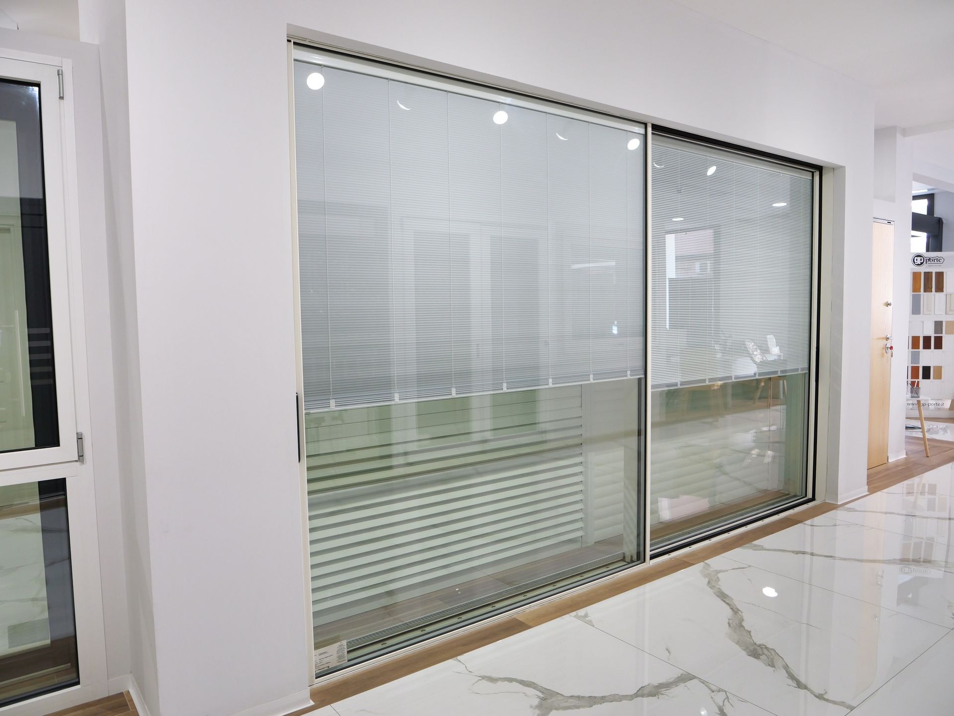 Una porta scorrevole in vetro con persiane orizzontali, inserita in una parete bianca con pavimento in marmo.