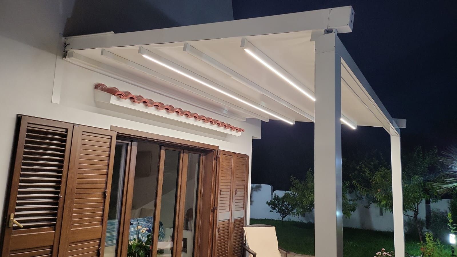 Un pergolato bianco con luci integrate, attaccato a una casa con persiane marroni, di notte.