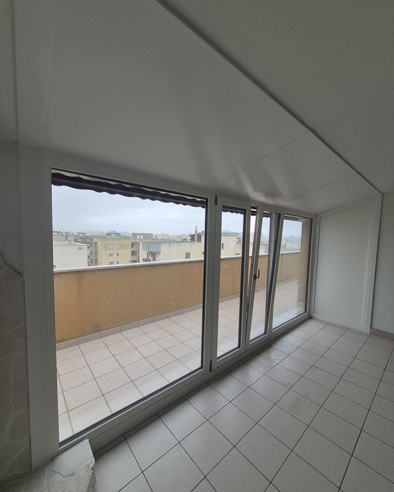 Vista interna della stanza con ampie finestre che si affacciano su un balcone cittadino. Pareti bianche, pavimento piastrellato.