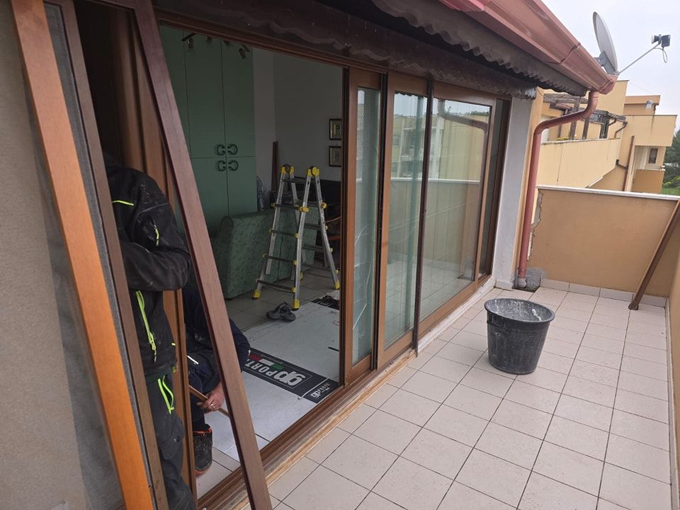 Persona che installa una porta scorrevole in vetro sul balcone. Telai marroni, secchio nero, piastrelle grigie.