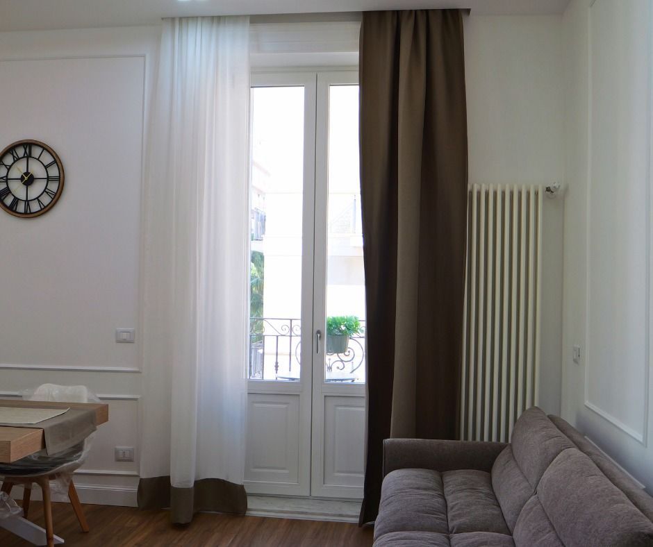 Soggiorno con tende bianche e marroni trasparenti, finestra, divano, termosifone e orologio.