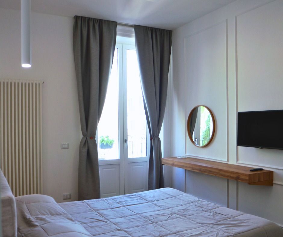 Camera da letto con letto, finestra, tende grigie, specchio e TV a parete.