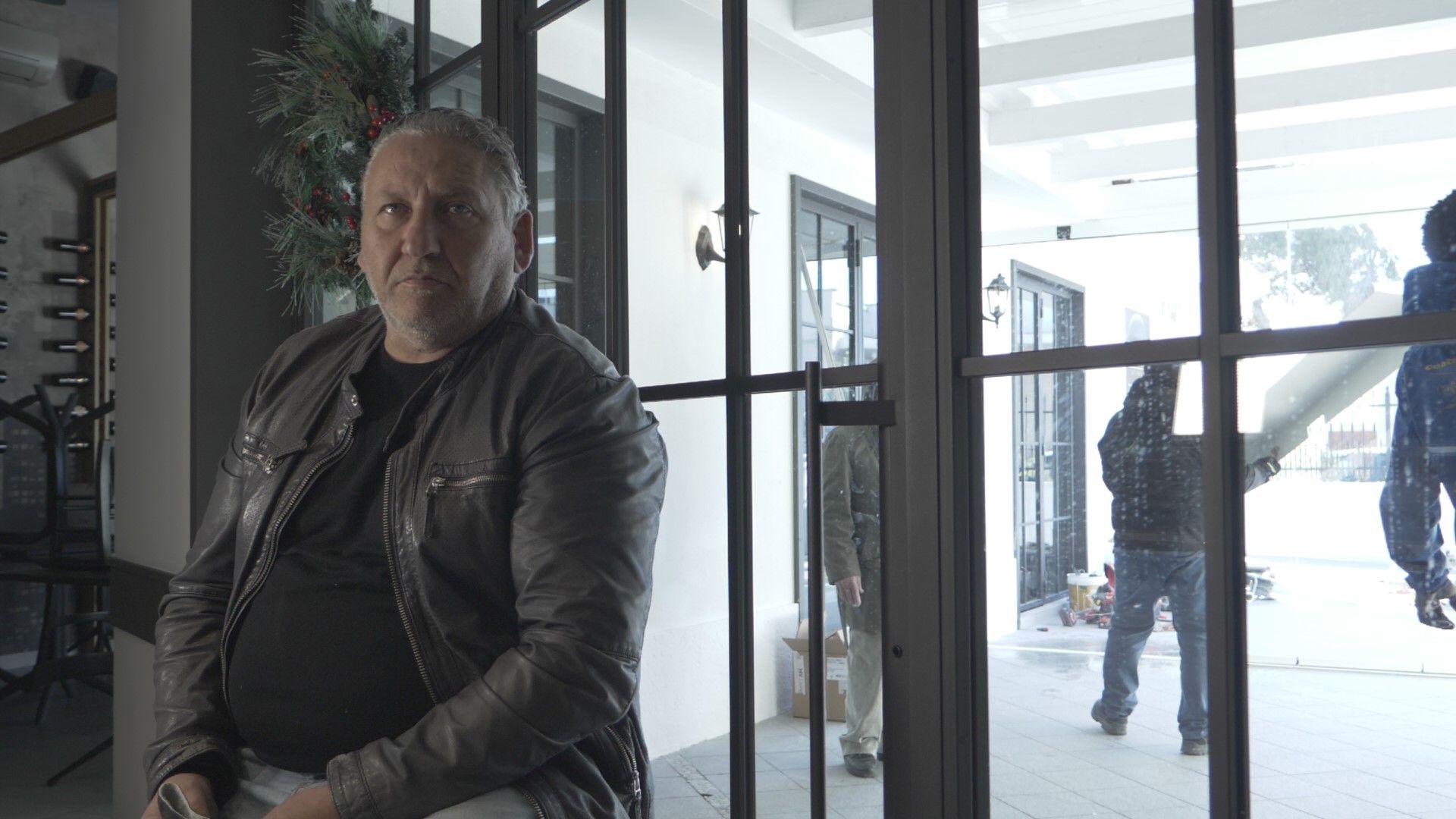 Uomo con giacca di pelle, guarda avanti, in casa vicino a porte a vetri, persone fuori che trasportano oggetti, neve.