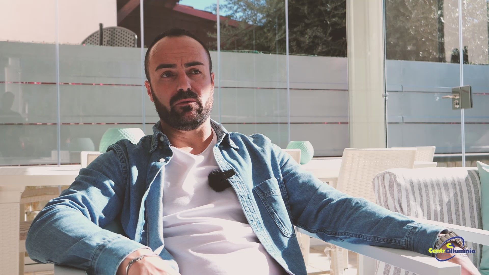 Uomo con la barba che indossa una camicia di jeans, seduto all'aperto, parla al microfono.