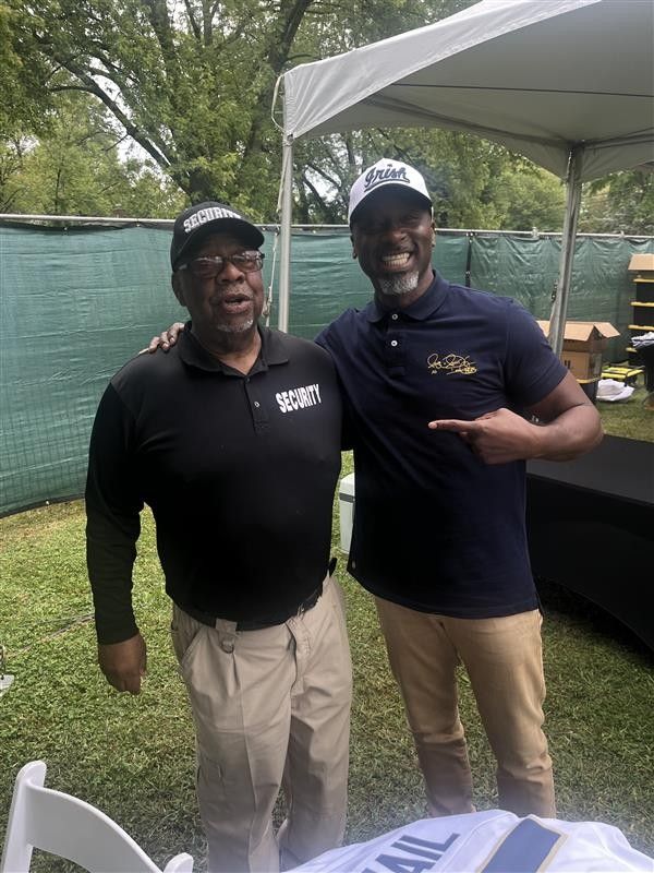 Two men smiling, posing for a photo under a tent. One points to the other, who has an arm around the first.