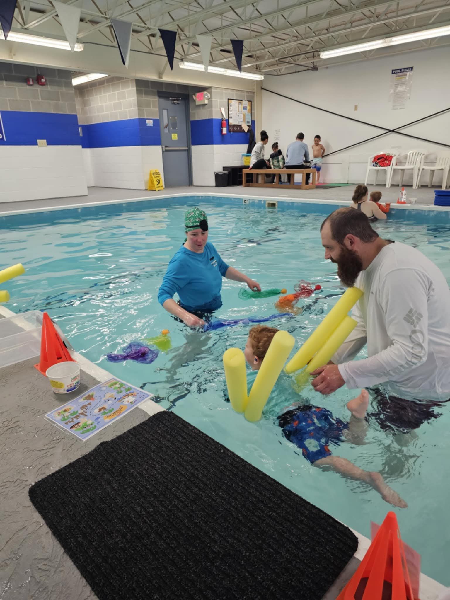 Indoor pool with two people using foam noodles during a water activity, with a coach nearby.