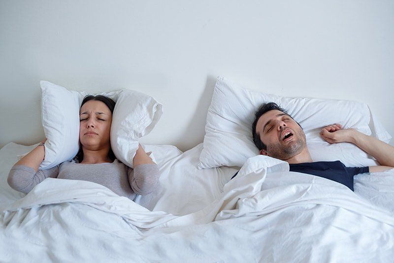 Man snoring in bed while his wife lays next to him, using a pillow to cover her ears
