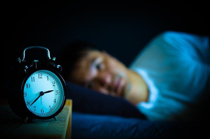 An alarm clock, with a blurred man laying in bed and staring at the alarm in the background