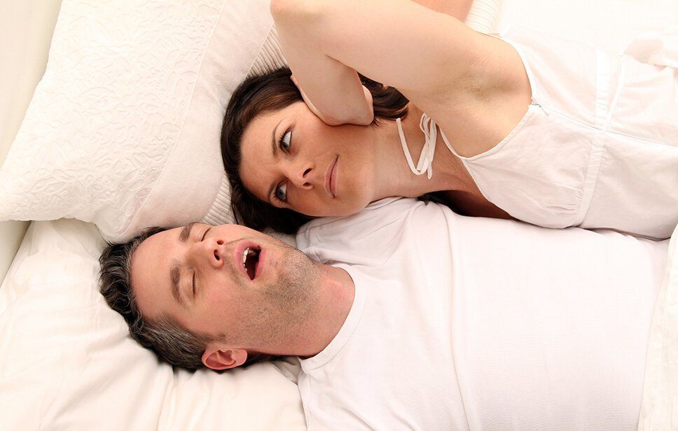 Man snoring while his wife lays next to him, looking angry and covering one of her ears
