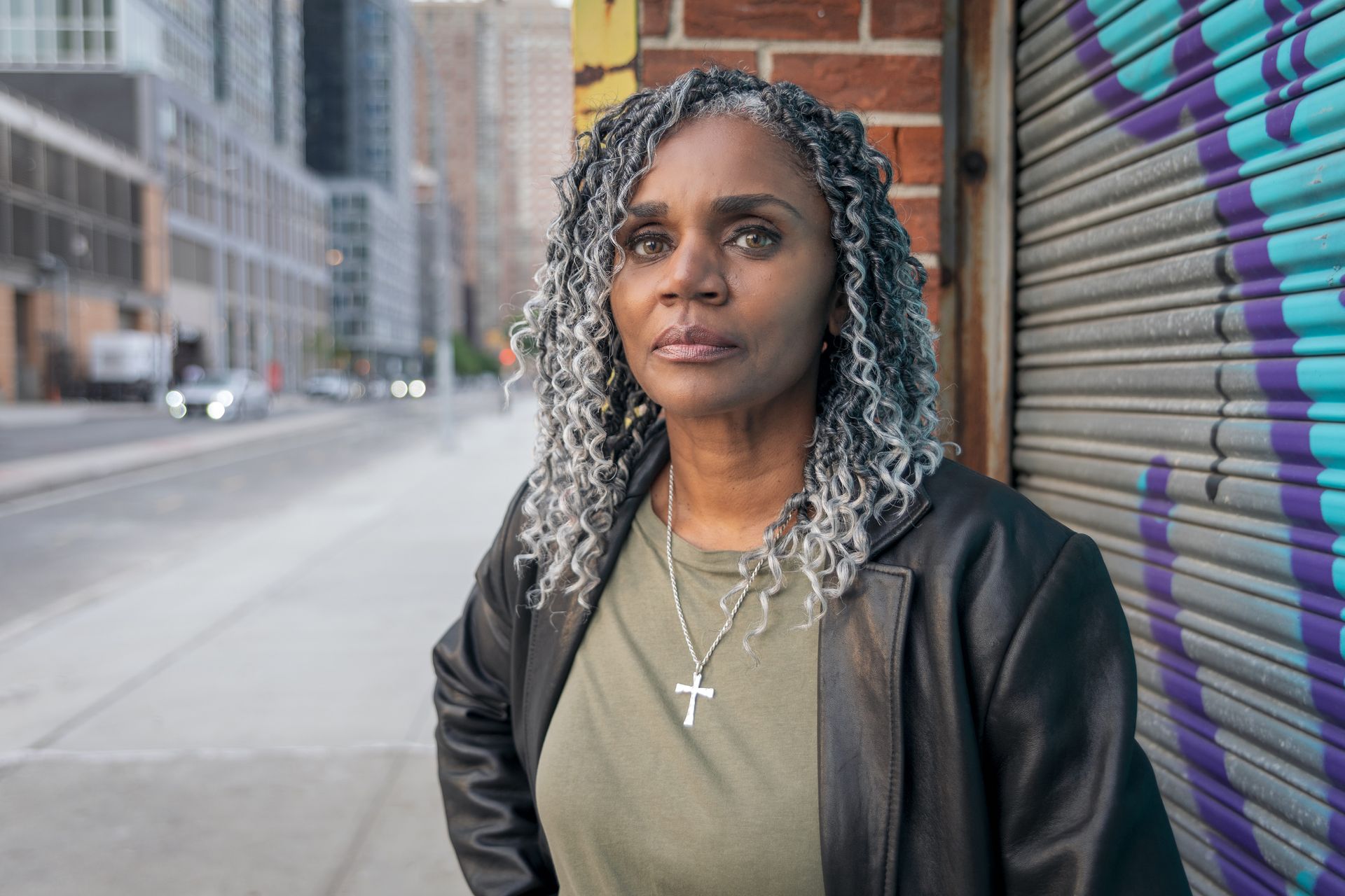 A woman with gray hair and a cross necklace is leaning against a brick wall.