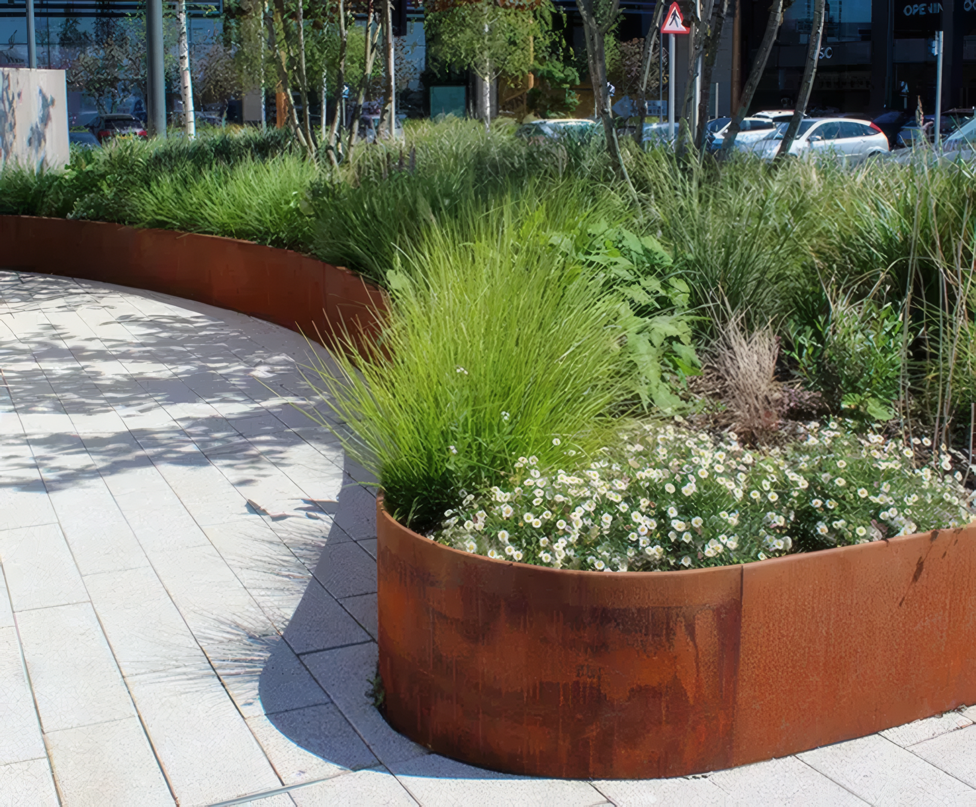 Curving rusted steel planter with various green plants and white flowers in a paved outdoor space.