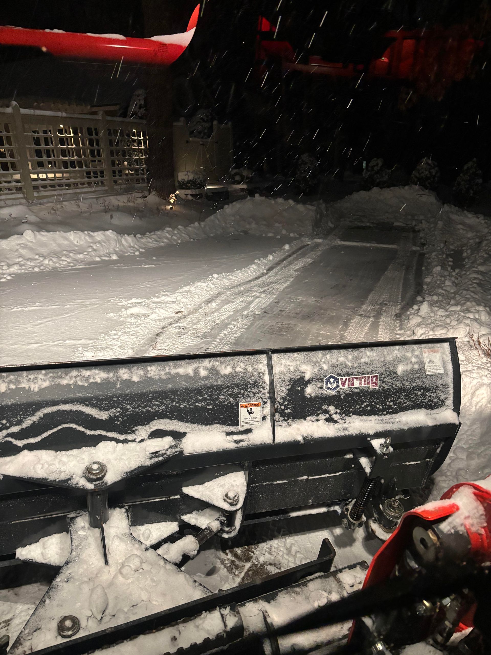 Snowplow clearing snow from a dark driveway at night, showing the plow blade and fresh tracks.