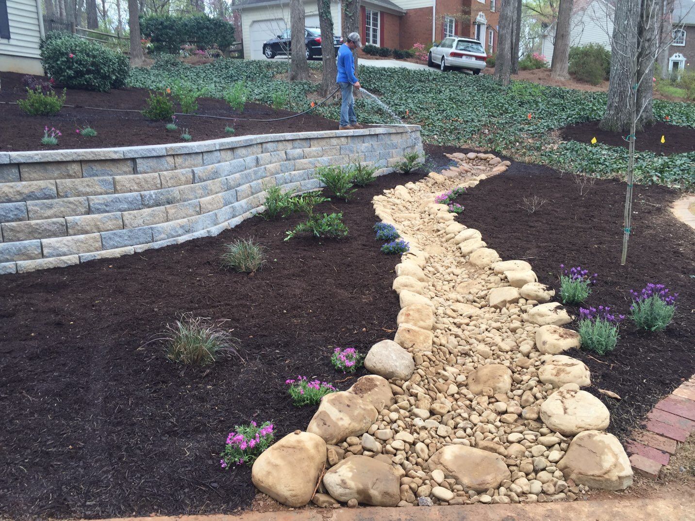 A landscaped yard with a stone retaining wall, mulch, and a dry creek bed; a person rakes in the background.