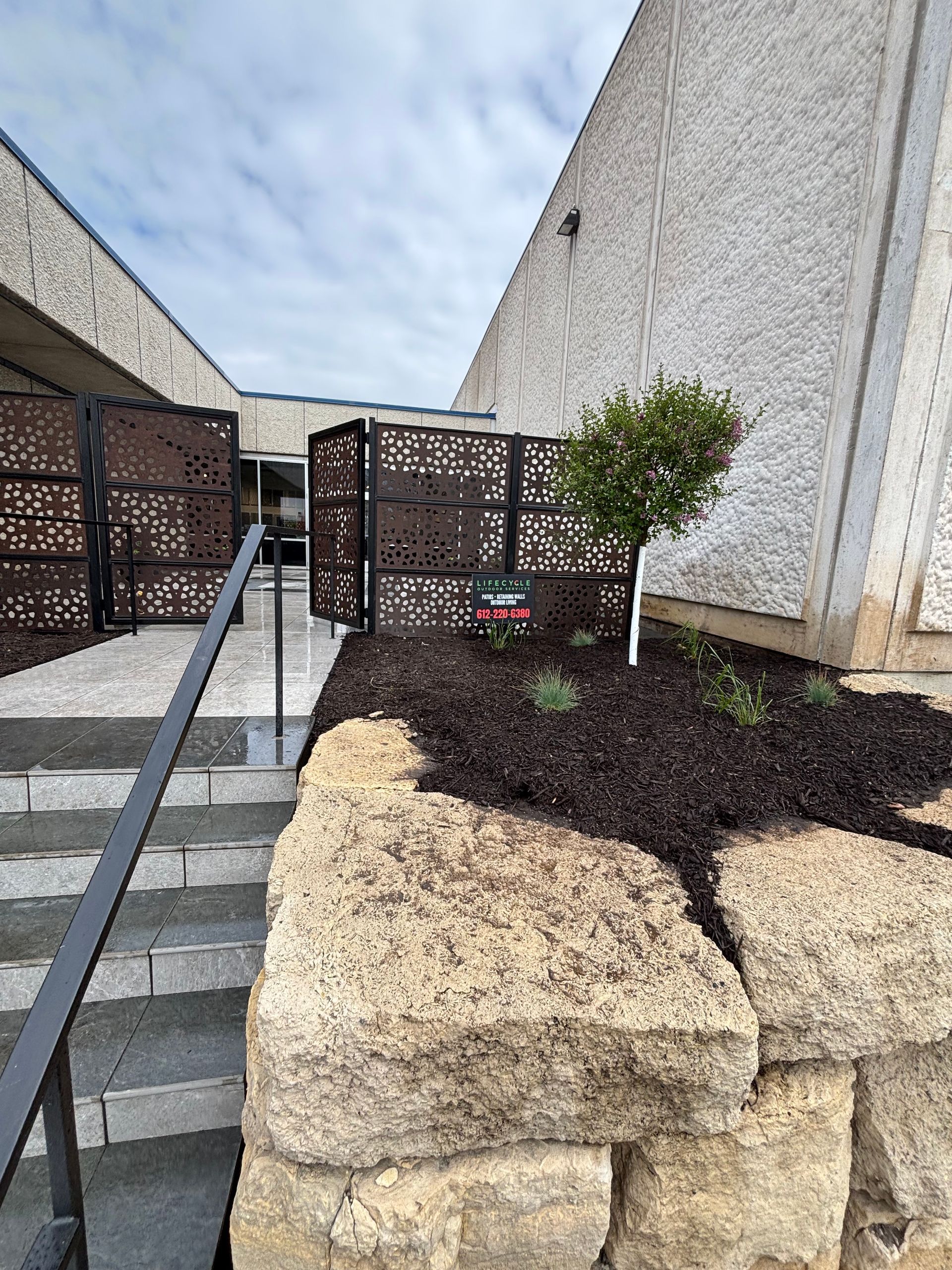 Entrance with stairs, decorative metal screens, and landscaping. Building has a textured, light-colored exterior.