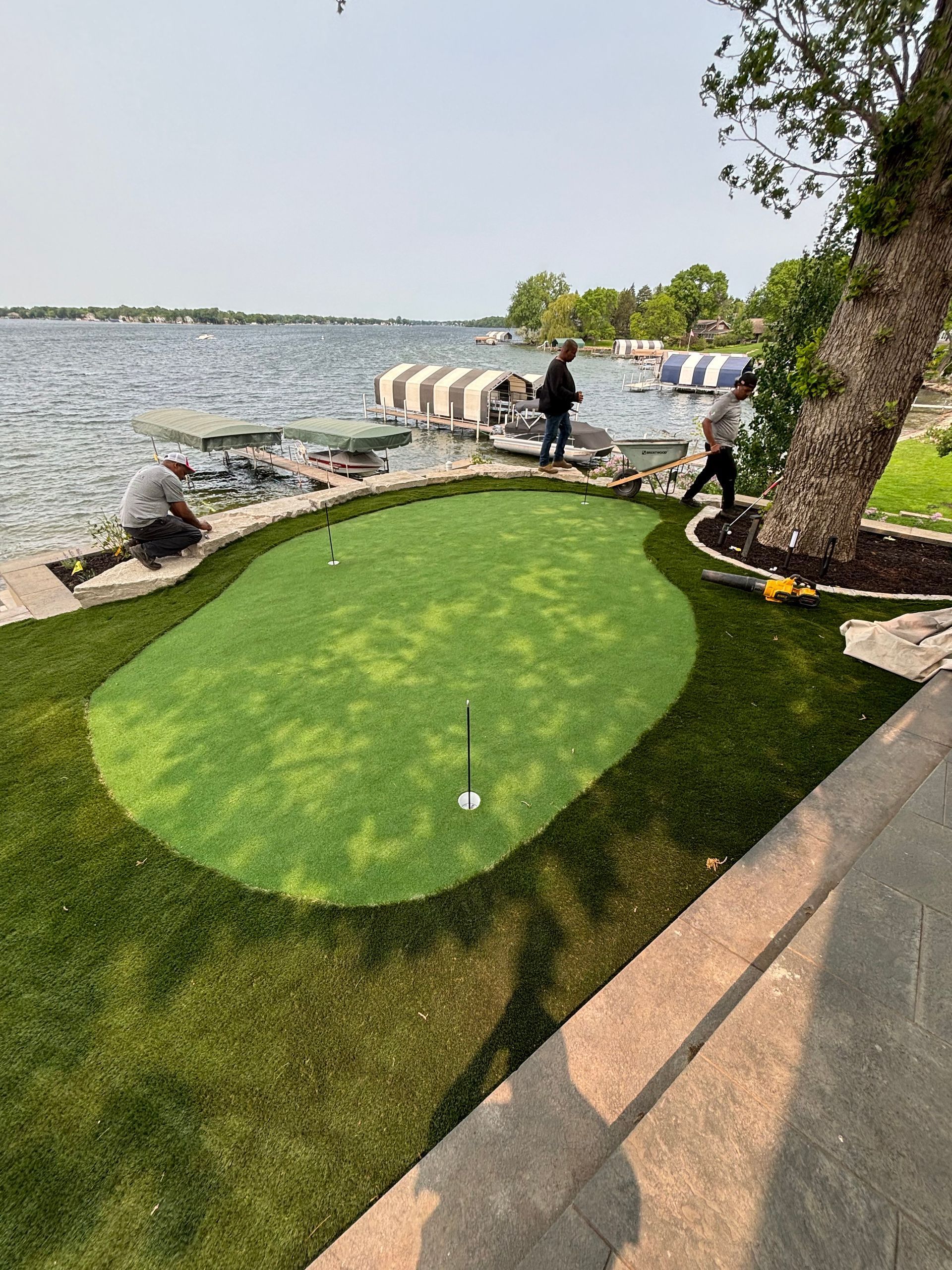 A putting green by a lake; two people near it. Green grass, blue water, sunny day.