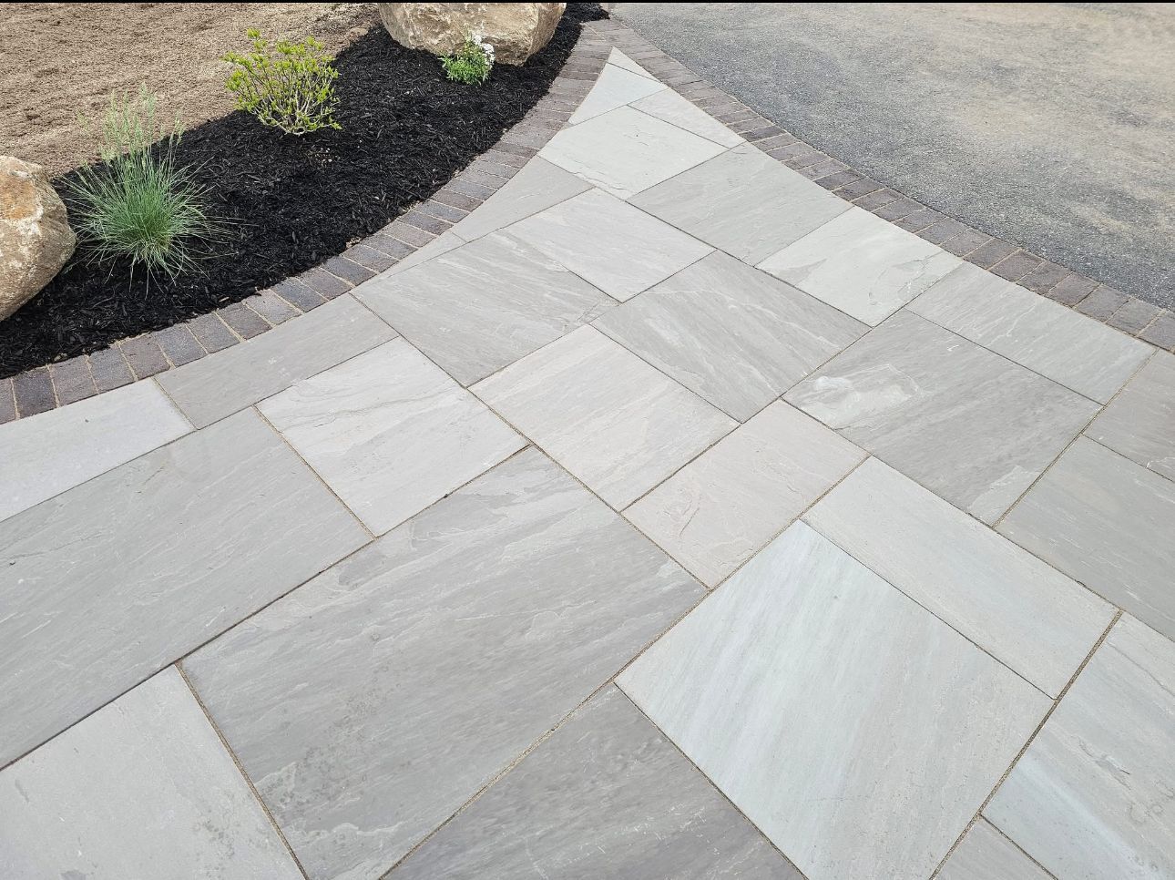 Gray sandstone patio with a black mulch border, next to gravel and some greenery.