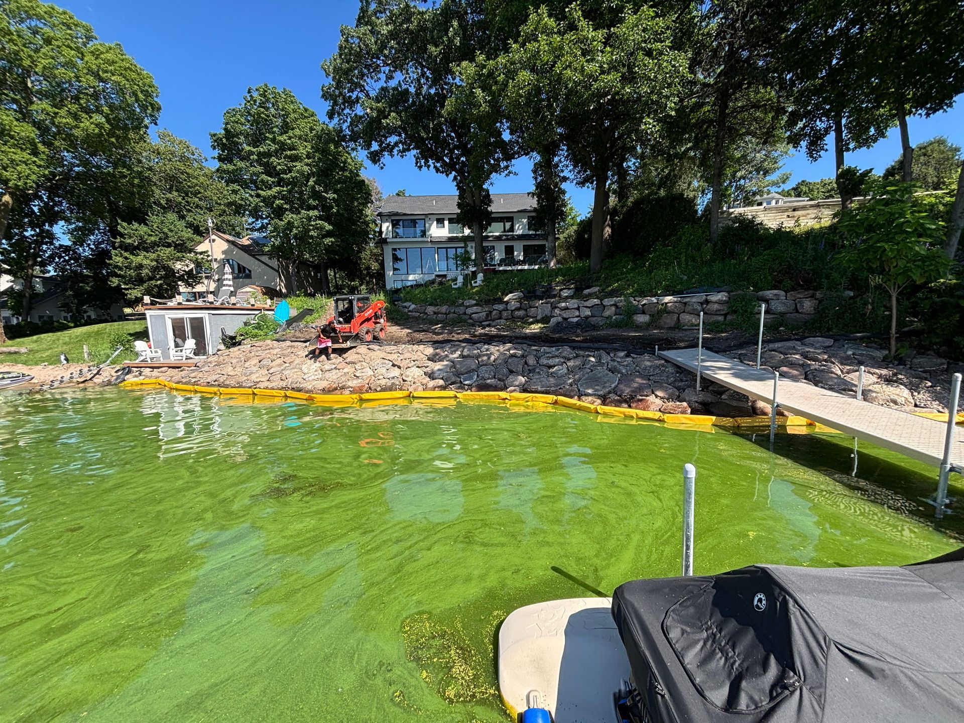 A construction site on a lakefront: a backhoe, rocks, dock, green water, houses, and trees.